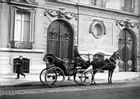 Femme cocher et son taxi hippomobile devant deux portes cocheres de la fin du XVIIIeme siècle. Paris, vers 1900